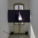 In einem schmalen Ausstellungsraum ist ein Multimedia-Kunstwerk zu sehen. Auf einem Bildschirm läuft ein Videospiel, und davor steht ein Mikrofon.