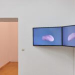 Im Ausstellungsraum sind zwei Kunstwerke zu sehen: Ein Videokunstwerk auf zwei BIldschirmen und im Raum dahinter ist eine eine Skulptur an der Wand angebracht.