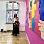 Ximena Ferrer Pizarro steht im Ausstellungsraum vor zwei Malereien, die an einer pinken Wand hängen. Sie spricht und gestikuliert.