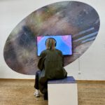 Eine Person sitzt vor einem Video-Kunstwerk auf einem Bildschirm in der Ausstellung. Sie trägt Kopfhörer und schaut auf den bildschirm.