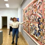 Die Künstlerin Lillian Morrissey steht im Ausstellungsraum vor Ihrem Kunstwerk, einem bunten Wandteppich und zeigt darauf.