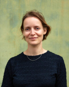 Porträtfoto Lotta Bartoschewski. Sie steht vor einer grünen Wand und schaut lächelnd in die Kamera.