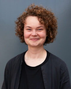 Porträtfoto von Ulrike Riebel. Sie steht vor einer grauen Wand und schaut lächelnd in die Kamera.