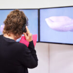 Eine Person steht im Ausstellungsraum vor zwei Bildschirmen. Sie trägt Kopfhörer und betrachtet ein Video auf den Bildschirmen.