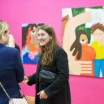 Zwei Personen stehen im Ausstellungsraum vor zwei Gemälden an einer pinken Wand und unterhalten sich.