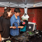 Zwei Personen stehen hinter einem DJ-Pult. Eine hält eine Schallplatte in der Hand.