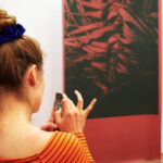 Eine Person steht vor einem Kunstwerk auf einem Vorhang und macht ein Foto davon mit ihrem Handy.
