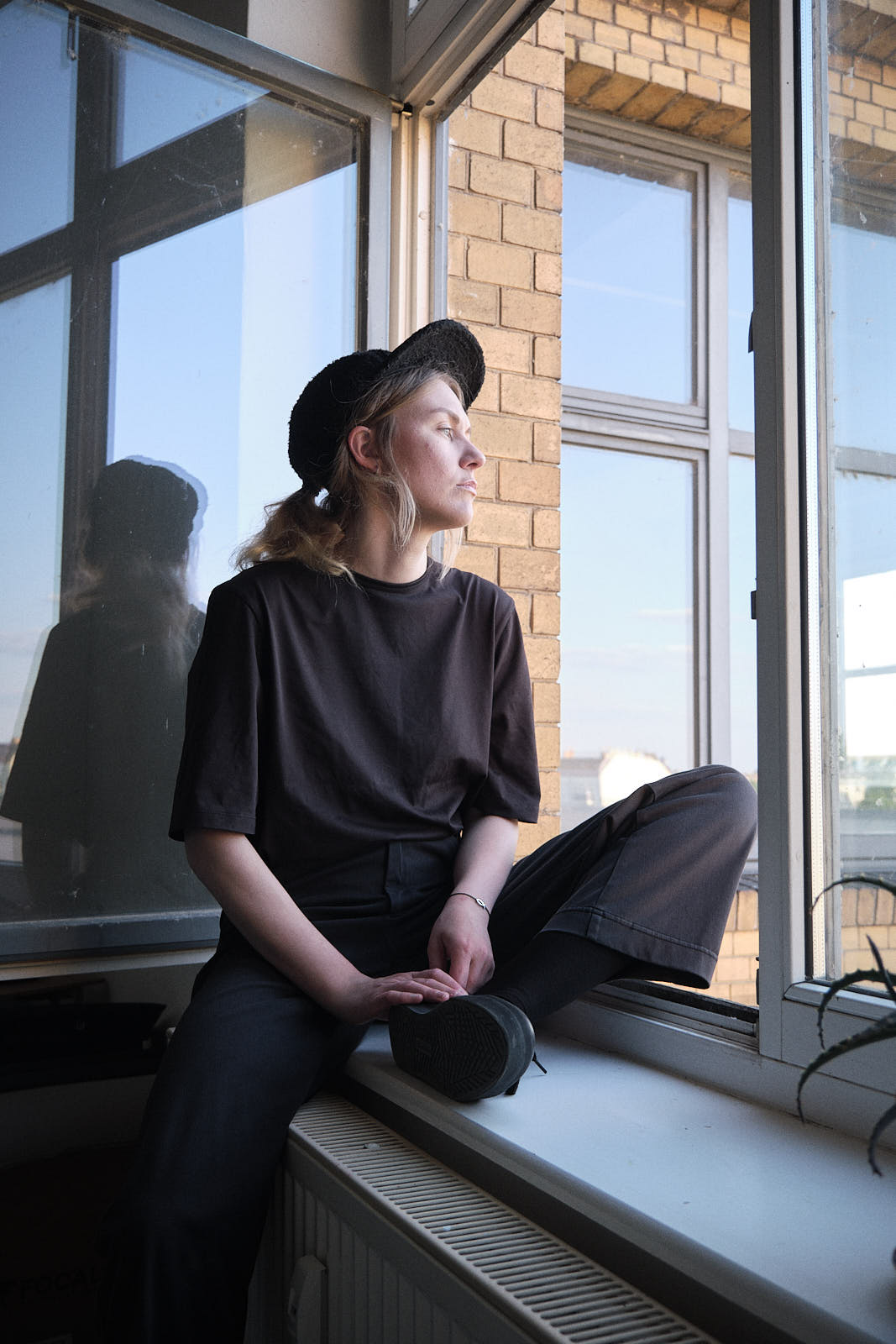 Porträtfoto von Bea Targosz. Die Künstlerin sitzt auf einem Fensterbrett und schaut aus dem Fenster. Sie trägt eine Mütze.