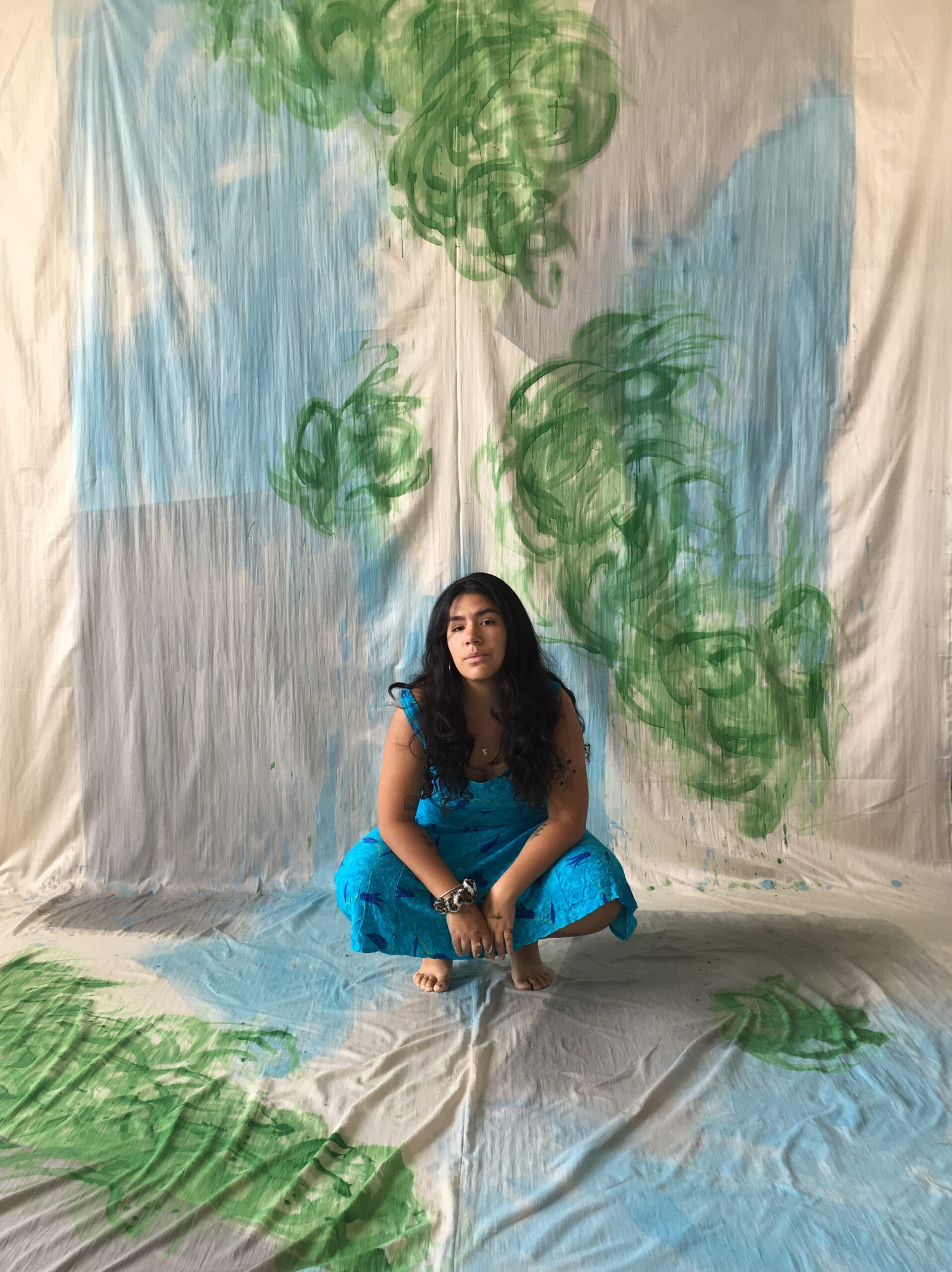 Porträtfoto von Ximena Ferrer Pizarro. Die Künstlerin sitzt in der Hocke vor einer bemalten Stoffbahn und schaut in die Kamera.